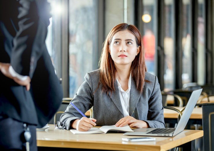 Upsetting face expression of young female employee sitting and writing note on table with laptop in front of angry manager